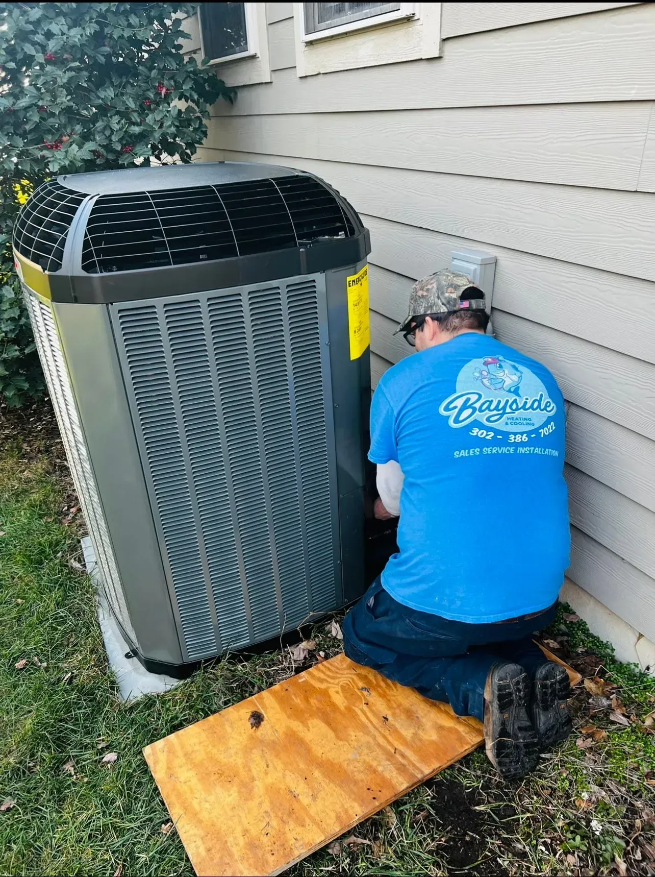 Worker kneeling beside outdoor air conditioner unit, making repairs with tools and plywood nearby