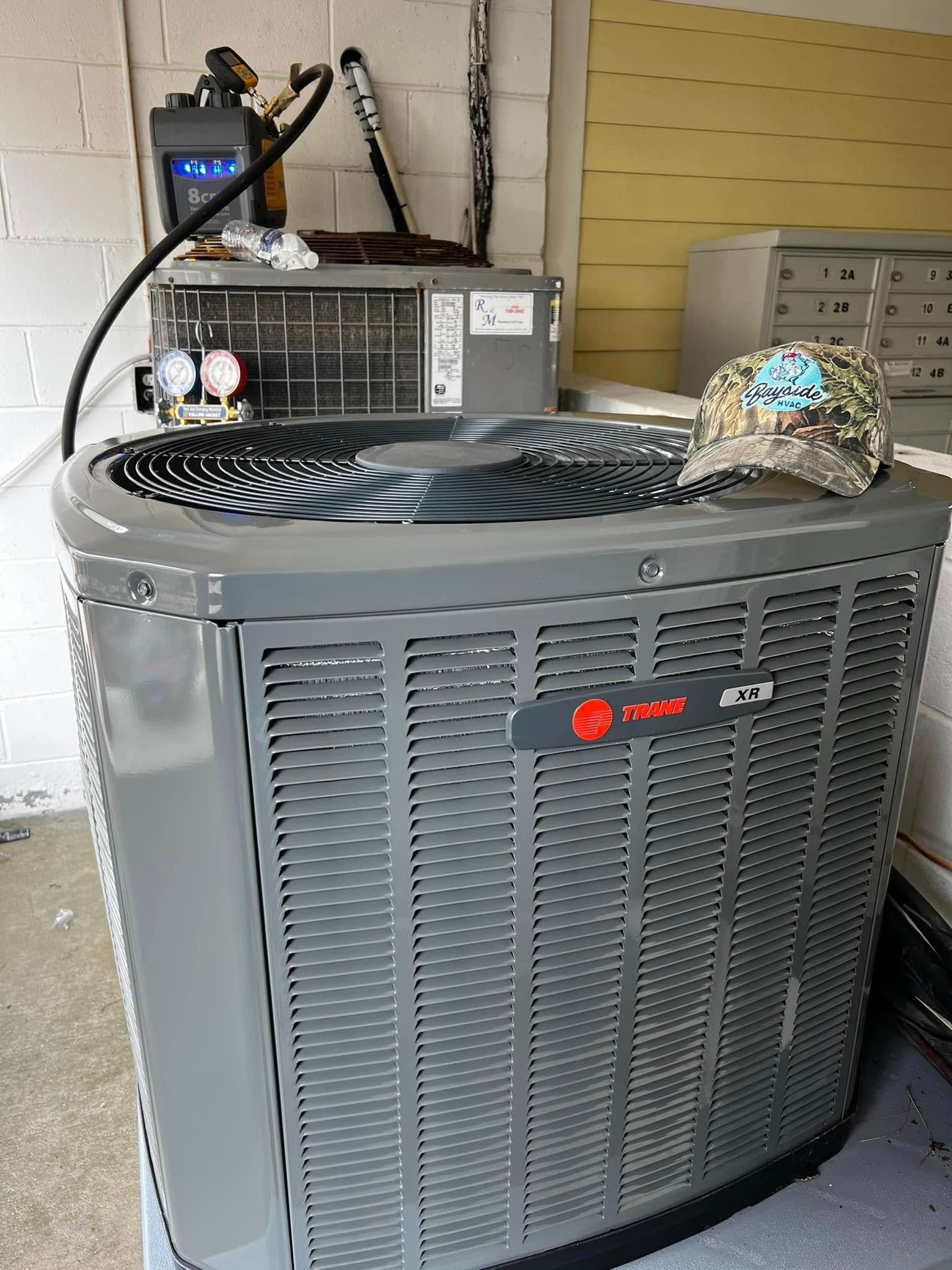 Gray outdoor air conditioner unit with a fan on top, set against a wall with equipment behind it
