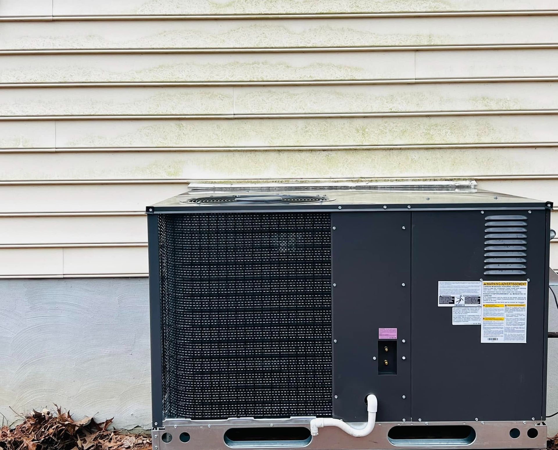 Outdoor HVAC condenser unit against a beige house wall with white siding and warning labels