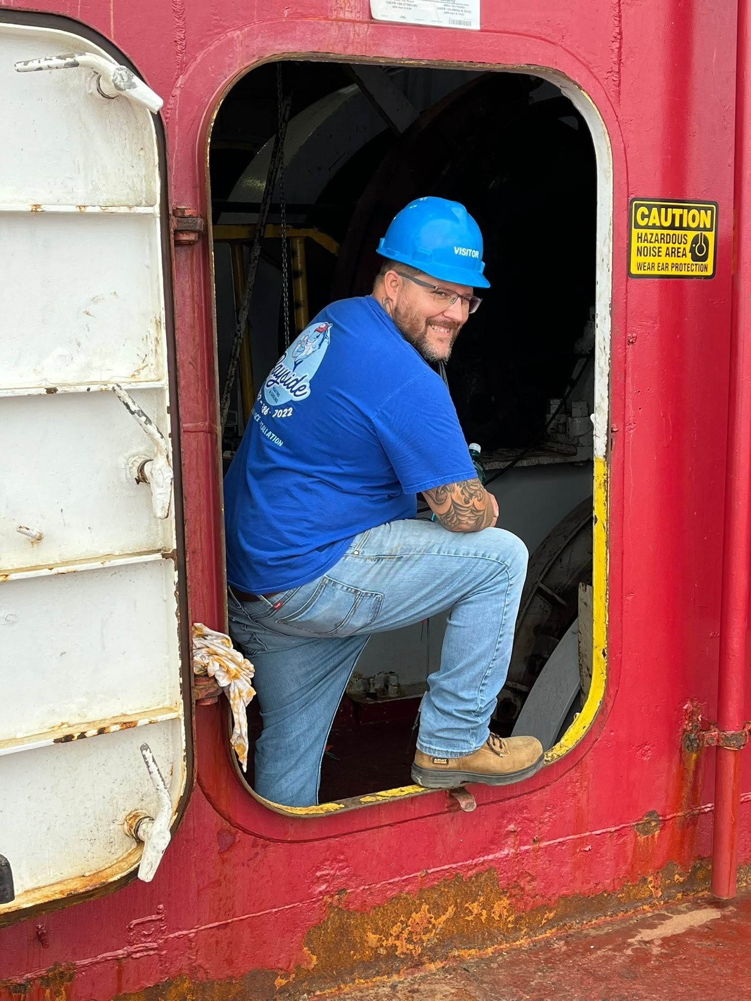 Worker in blue hard hat crouches in a red industrial hatch opening.