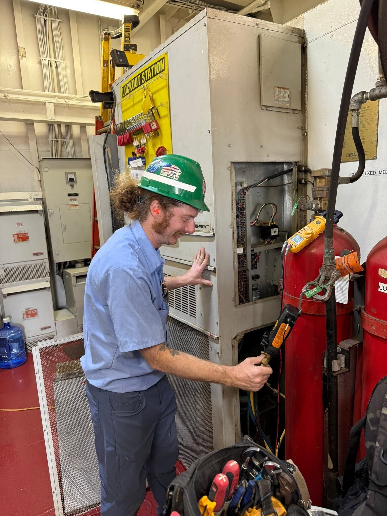 Worker in hard hat adjusting equipment in a utility room beside fire extinguishers.