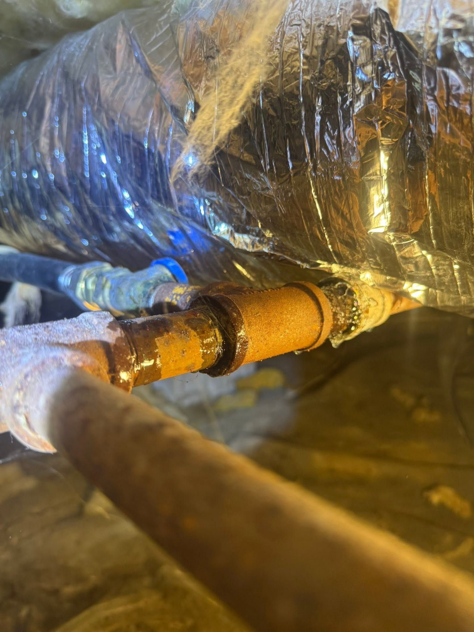 Gloved hands inspect a rusted pipe wrapped in insulation and plastic sheeting in a dim industrial space