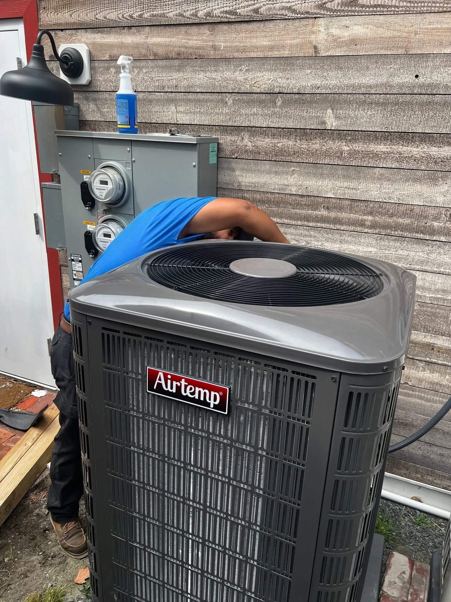 Air conditioning unit being serviced outdoors beside a wall, with technician and tools visible