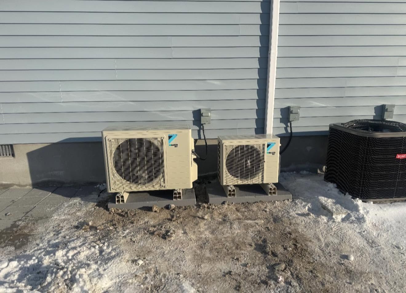Two outdoor HVAC condenser units beside a building wall on snowy ground