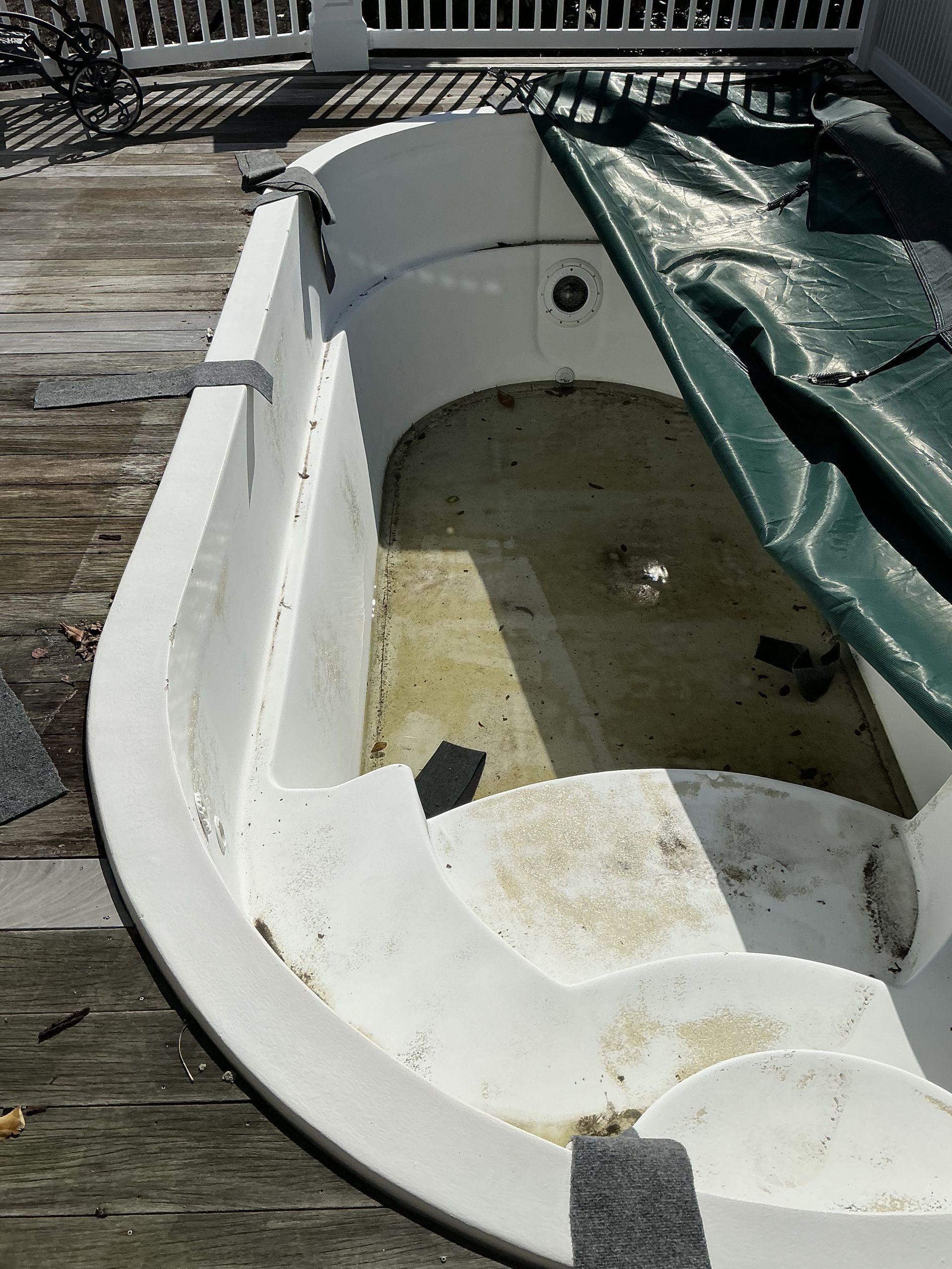 A dirty, empty white hot tub on a wooden deck, partially covered by a green tarp.