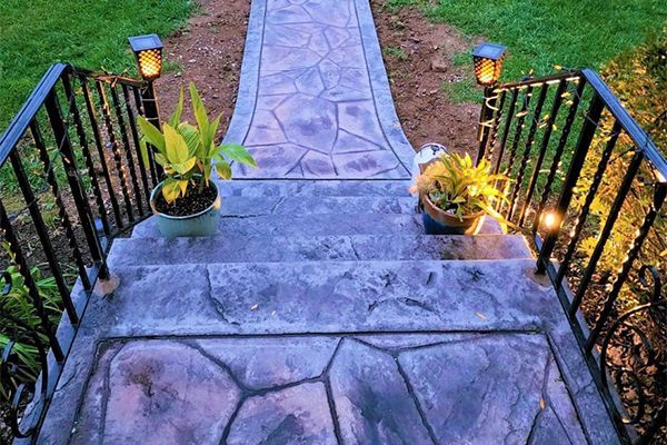 A stone walkway with stairs and a wrought iron railing is lit up at night.