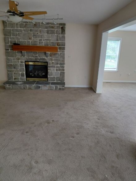 Living room with stone fireplace, beige carpet, and open doorway to another room with a window.