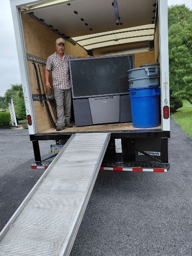 Man in truck, loading furniture: large TV, containers. Ramp extended, truck parked outside.
