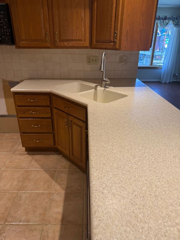 Kitchen with wood cabinets, off-white countertop, and a double sink. Tile flooring and a window in the background.