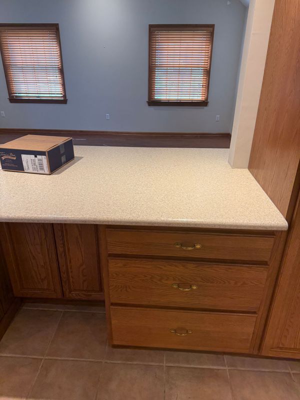 Oak kitchen cabinets with drawers, countertop, two windows with blinds, and a box on the counter.