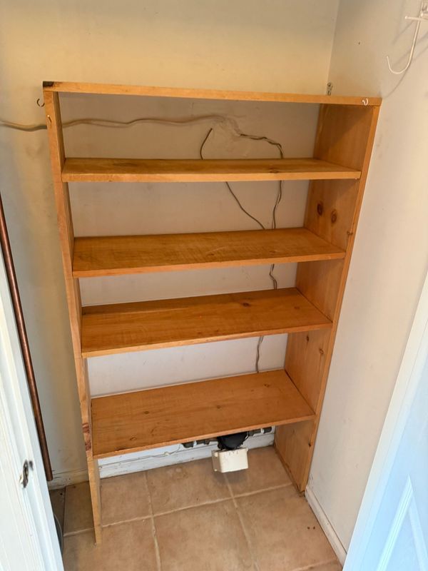 Wooden bookshelf with five shelves against a beige wall, a tan tile floor, and electrical wires.