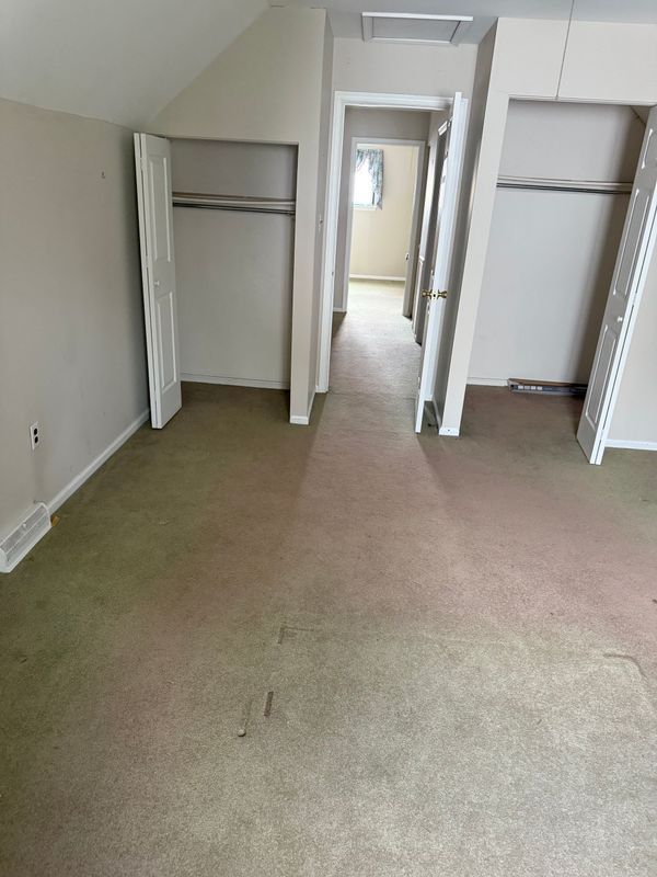 Bedroom with two closets, beige walls, worn green and pink carpet, doorway to another room.