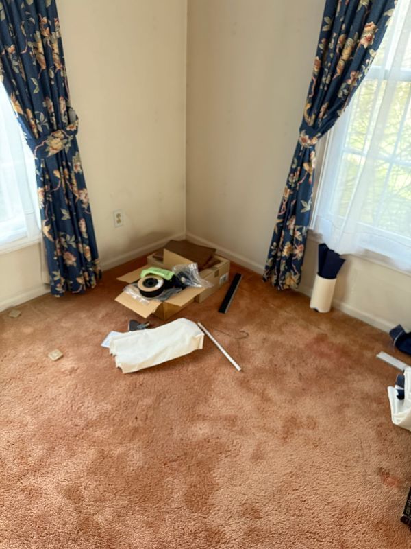 A room with dingy peach carpet, blue floral curtains, and a pile of debris on the floor near a window.