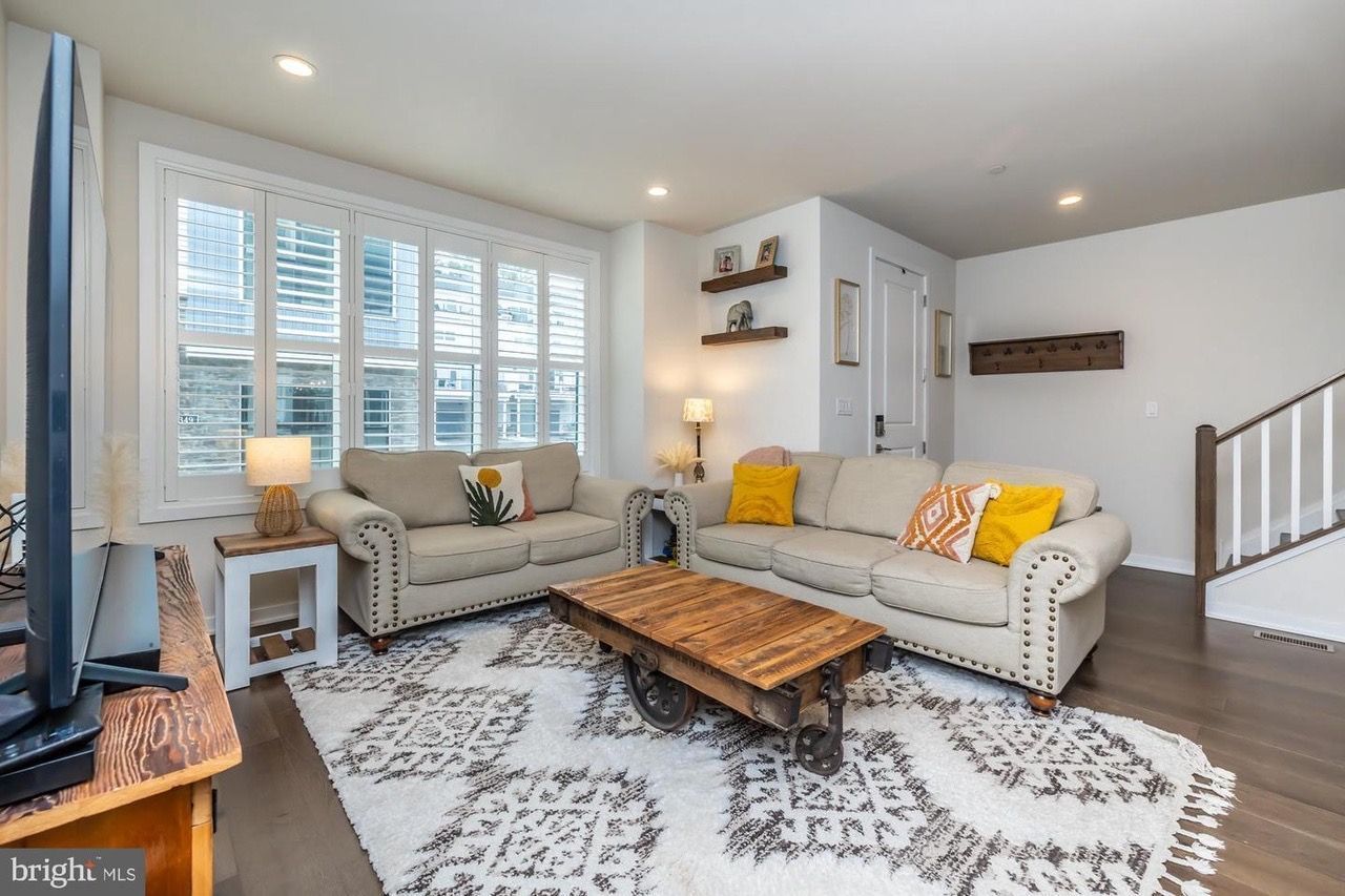 Living room with neutral-toned couches, patterned rug, wooden coffee table, and large window with shutters.