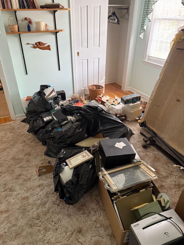 A room cluttered with black trash bags, boxes, and miscellaneous items on beige carpet. A door and window are visible.
