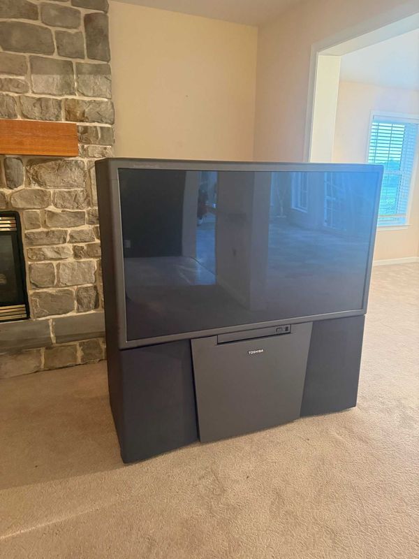 Large gray projection TV in a room with a stone fireplace and beige carpet.