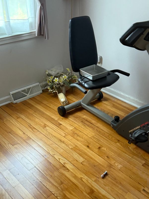 Recumbent exercise bike with a scale on the seat, next to a window and flowers on a wood floor.