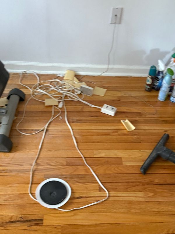 Tangled electrical cords and power strips on a wooden floor near a wall outlet and vacuum cleaner.