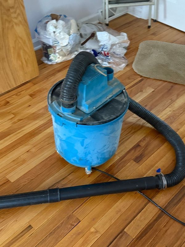 Blue vacuum cleaner on hardwood floor with black hose and debris in background.