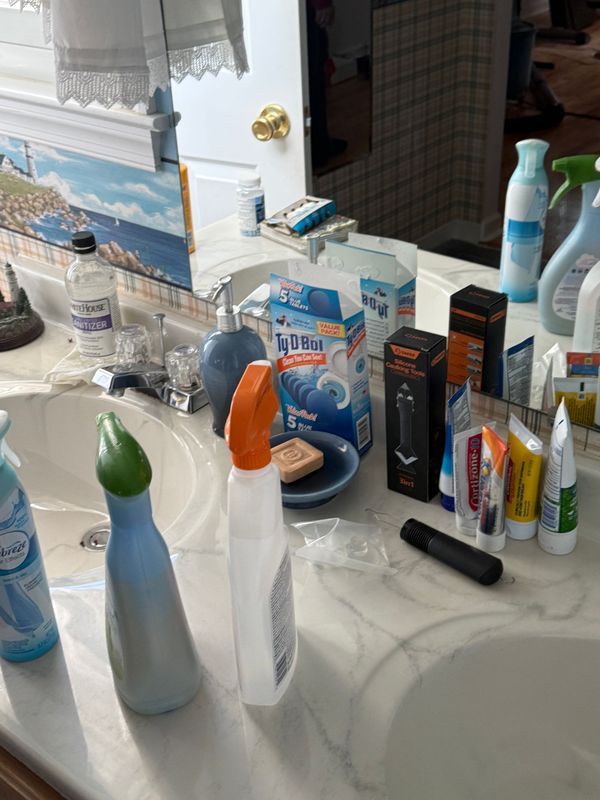Bathroom countertop cluttered with cleaning supplies and toiletries, reflecting in the mirror.