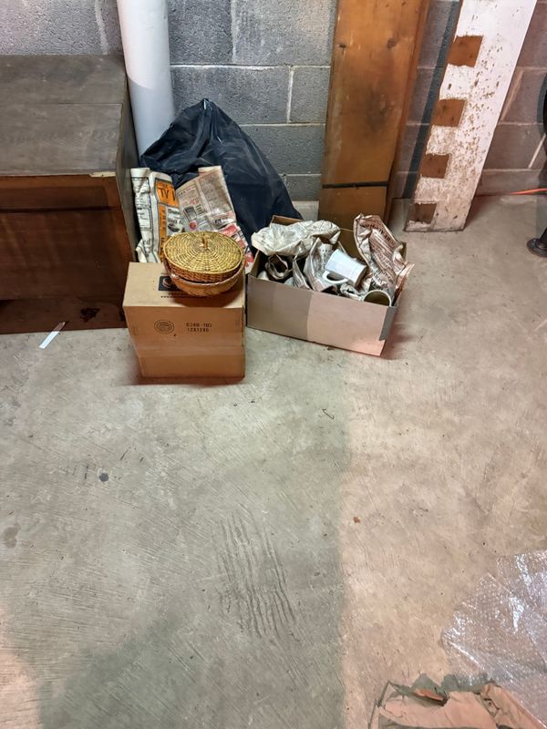 Boxes and items on a concrete floor in a basement setting. Includes a basket, dishes, and wood.