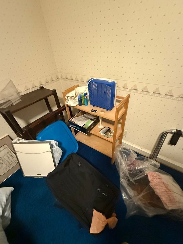 Cluttered room with a wooden shelf, furniture, blue carpet, and wallpaper with sailboat pattern.