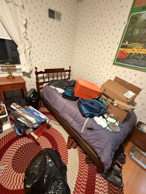 Cluttered bedroom with bed, luggage, boxes, and a rug. The walls have floral wallpaper, and there's a window.