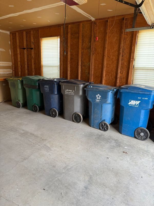 Seven wheeled trash bins of various colors lined up inside a garage.