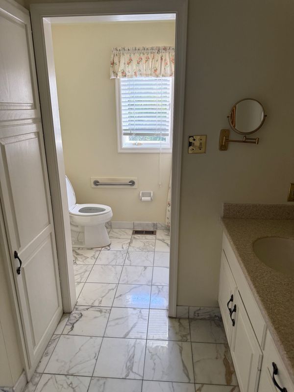 Bathroom interior with a toilet, vanity, and a window. White cabinets, light-colored walls, and tiled floor.