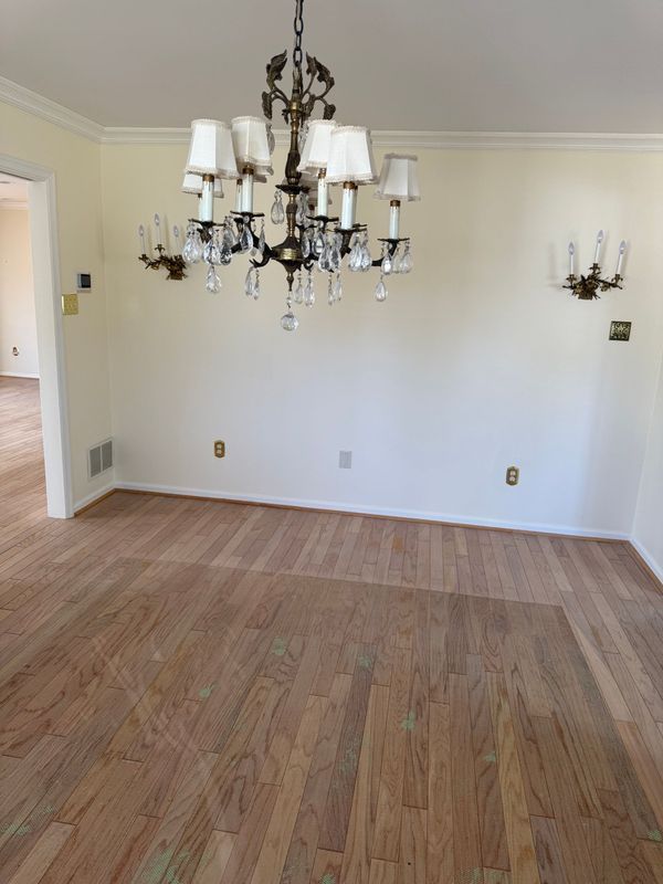 Empty dining room with chandelier and sconces, wood floor, off-white walls, and doorway to another room.