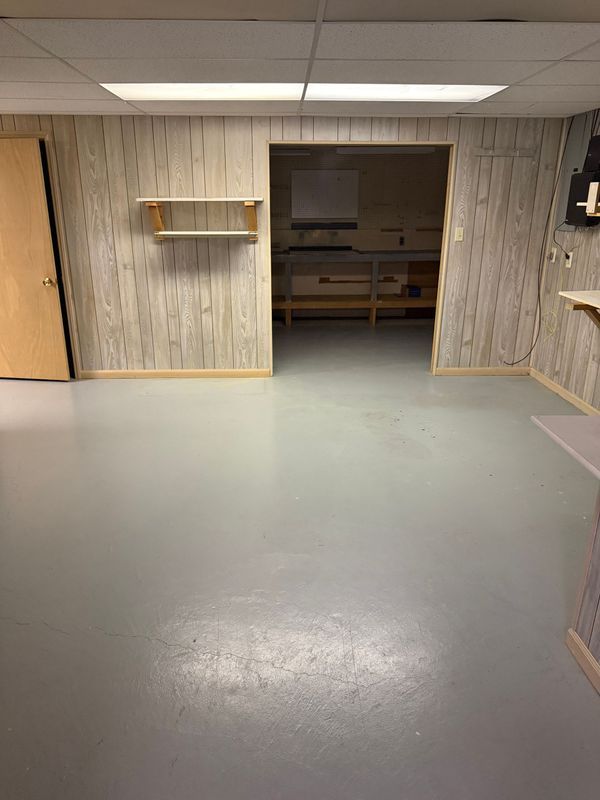 Empty room with light gray floor, light wood panel walls, open doorway leading to a workshop area.