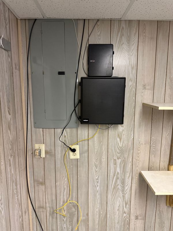 Electrical panel and network boxes mounted on a wood-paneled wall, near outlets and shelving.