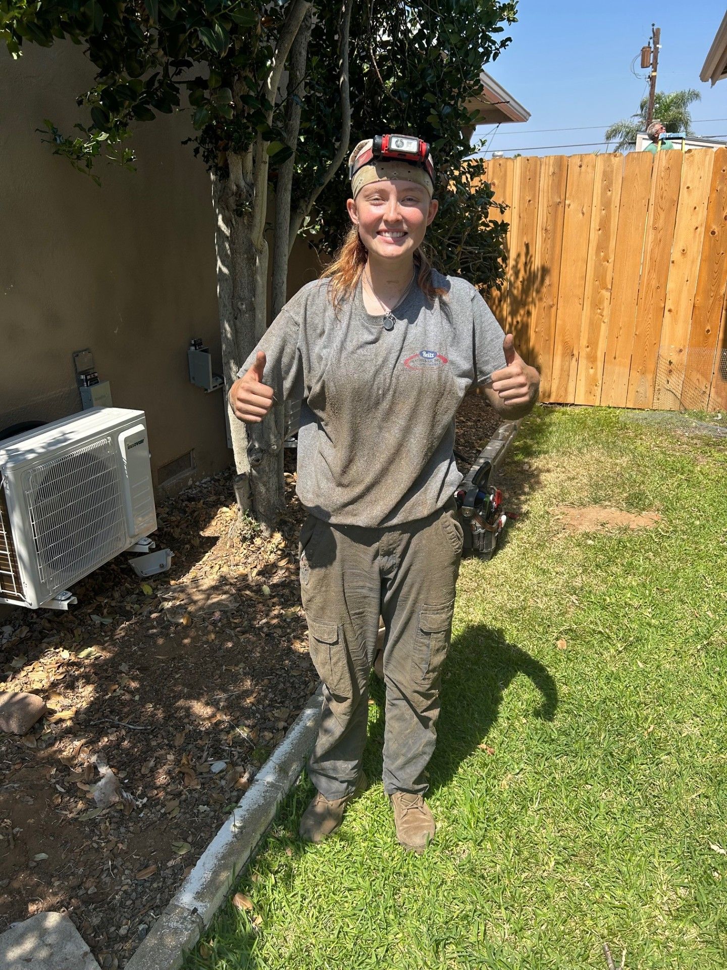 Woman covered in dirt, thumbs up in a backyard with AC unit and wooden fence.