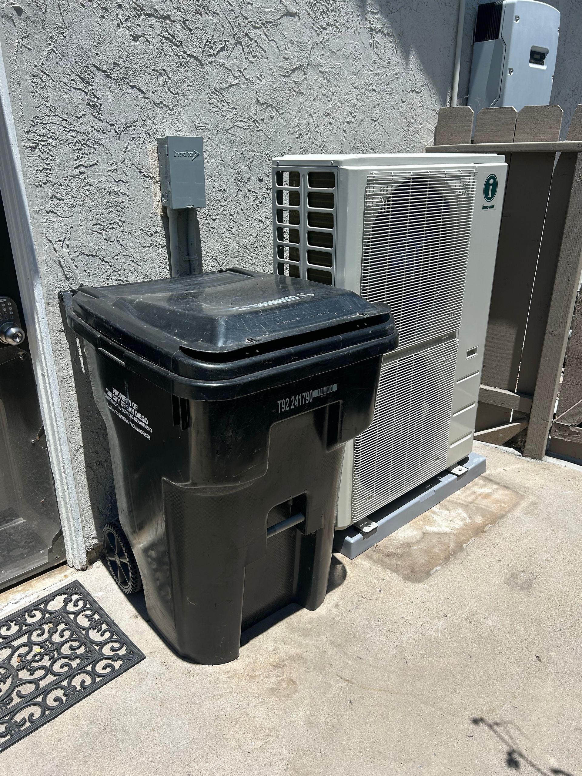 Black trash bin and air conditioning unit next to a wall. A small electrical box is mounted above the bin.