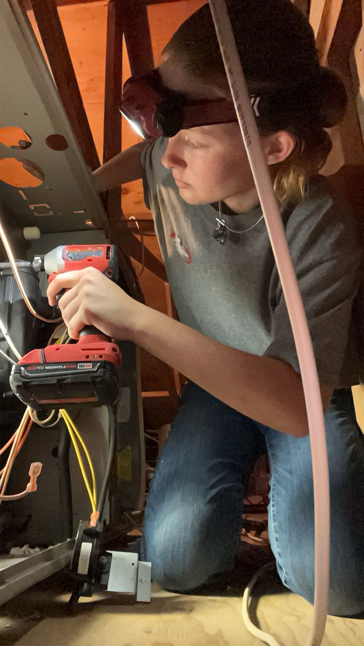 Person wearing a headlamp works on equipment in an attic, using a power drill. They are wearing jeans and a gray shirt.