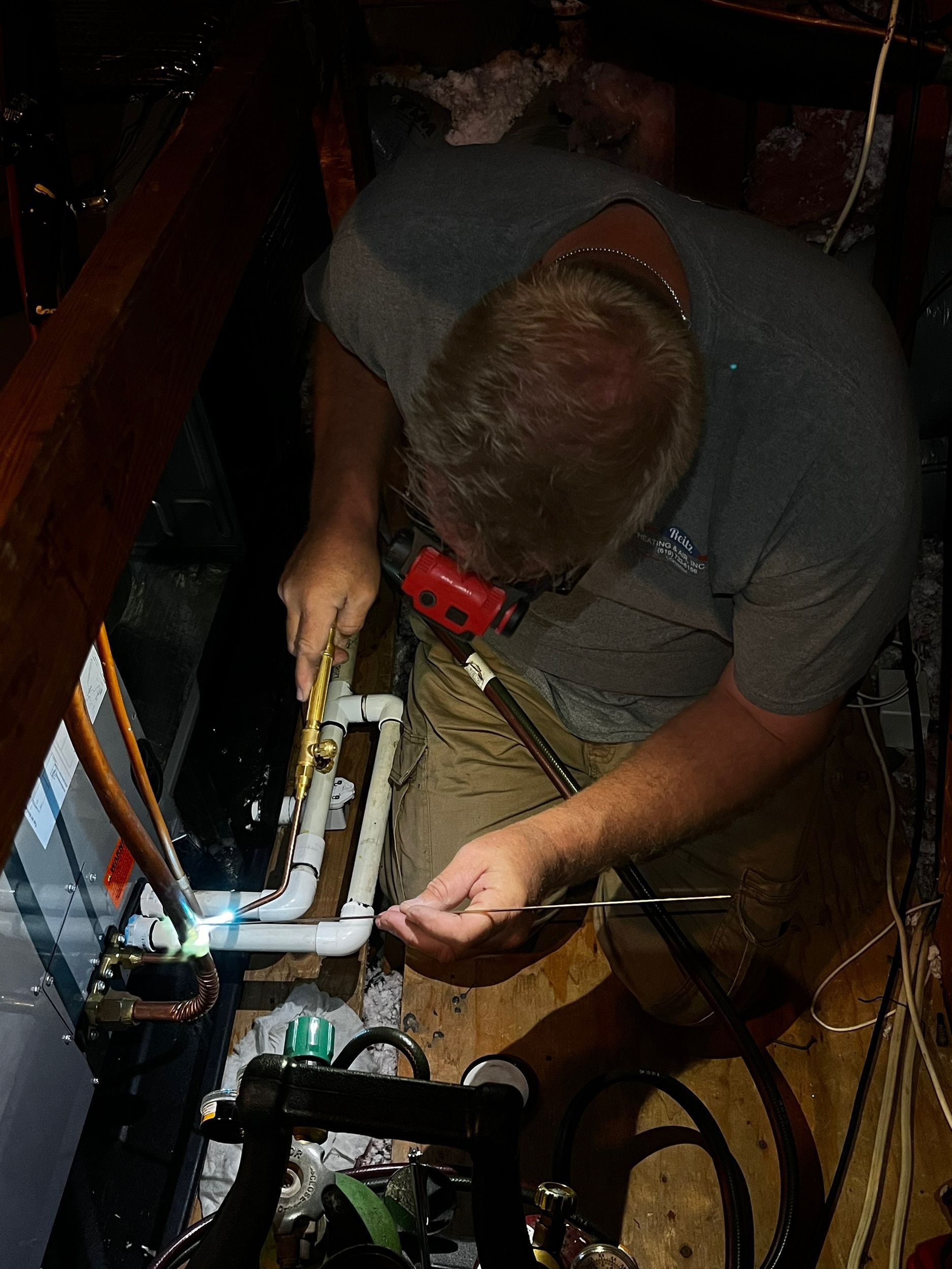 A man working on plumbing in a dimly lit attic, wearing safety glasses, cutting a white pipe with a saw.