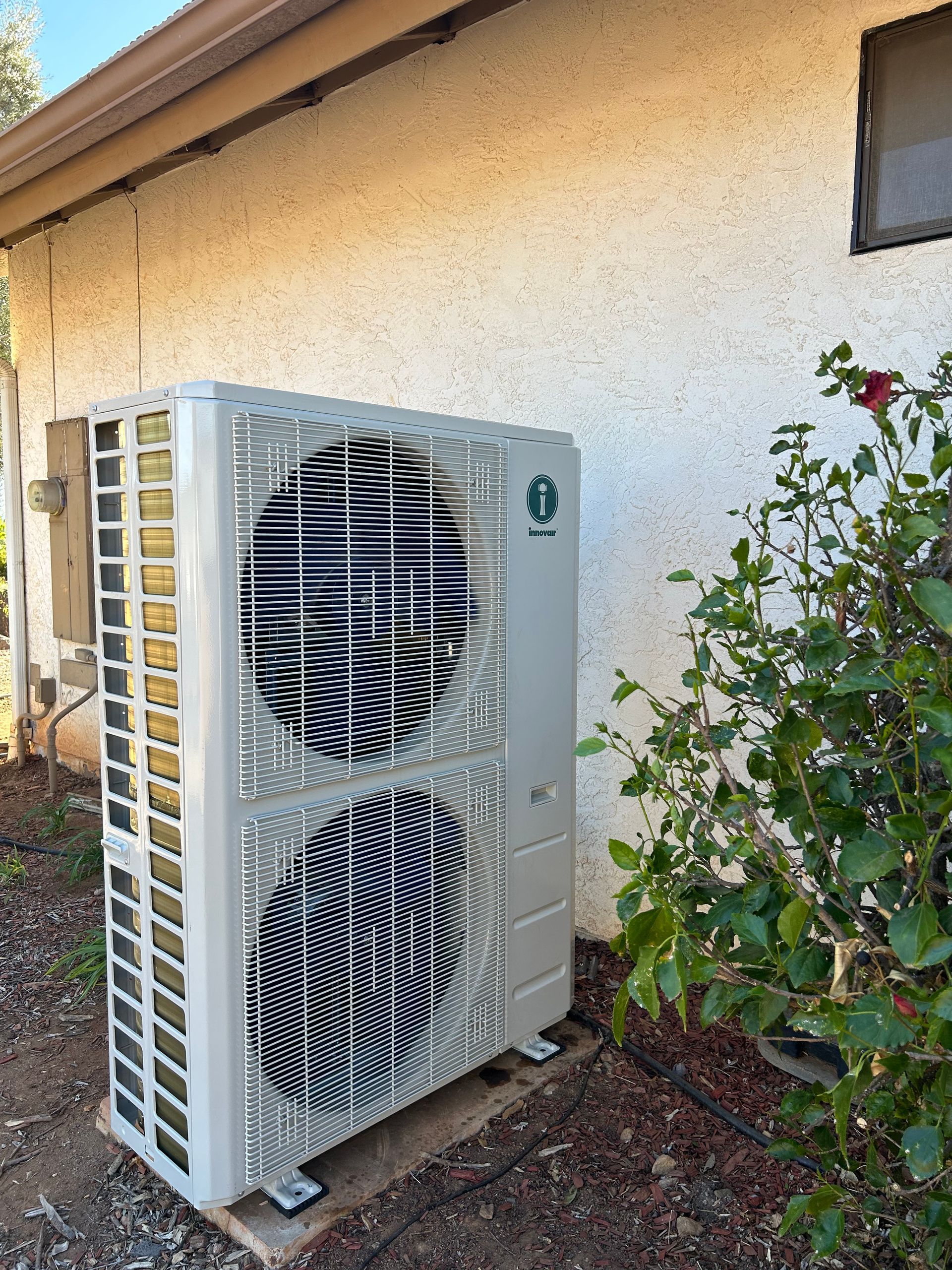 A silver heat pump unit next to a stucco wall, with foliage to the right.