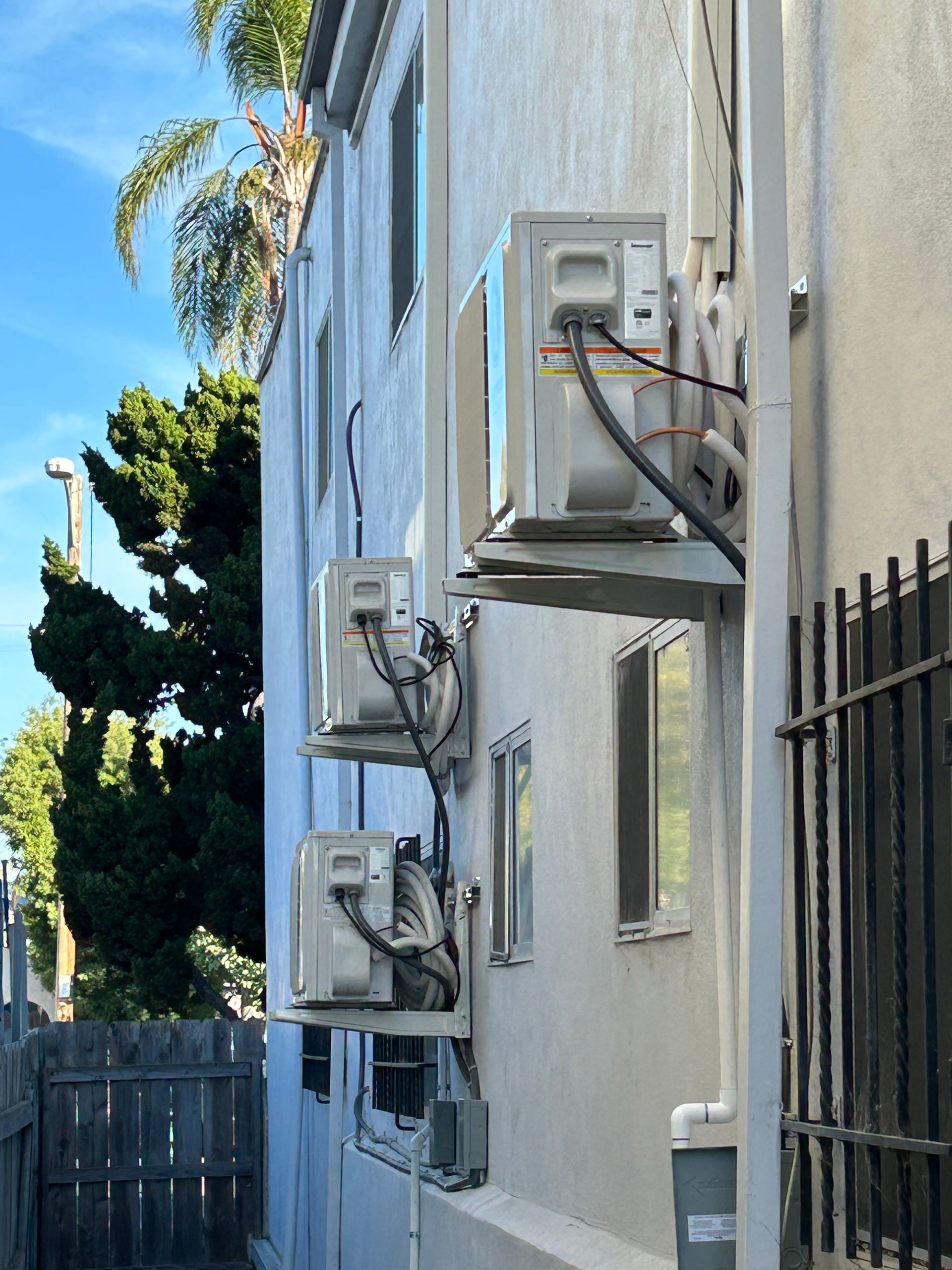 Three air conditioning units mounted on the exterior wall of a multi-story building. A fence and a tree are in the background.