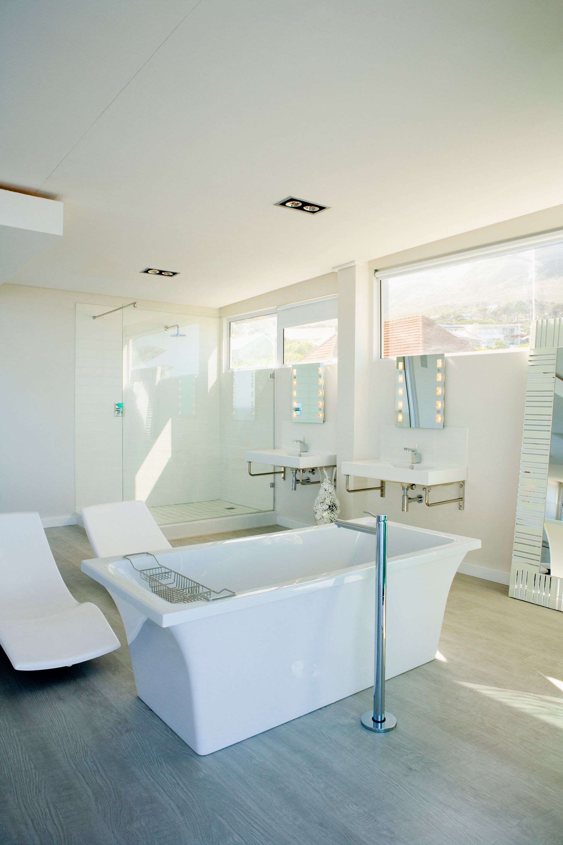 Bright white bathroom with a freestanding tub, two lounge chairs, and large windows.