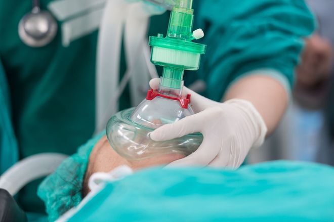 Person in operating room, receiving oxygen from a green mask held by gloved hands.