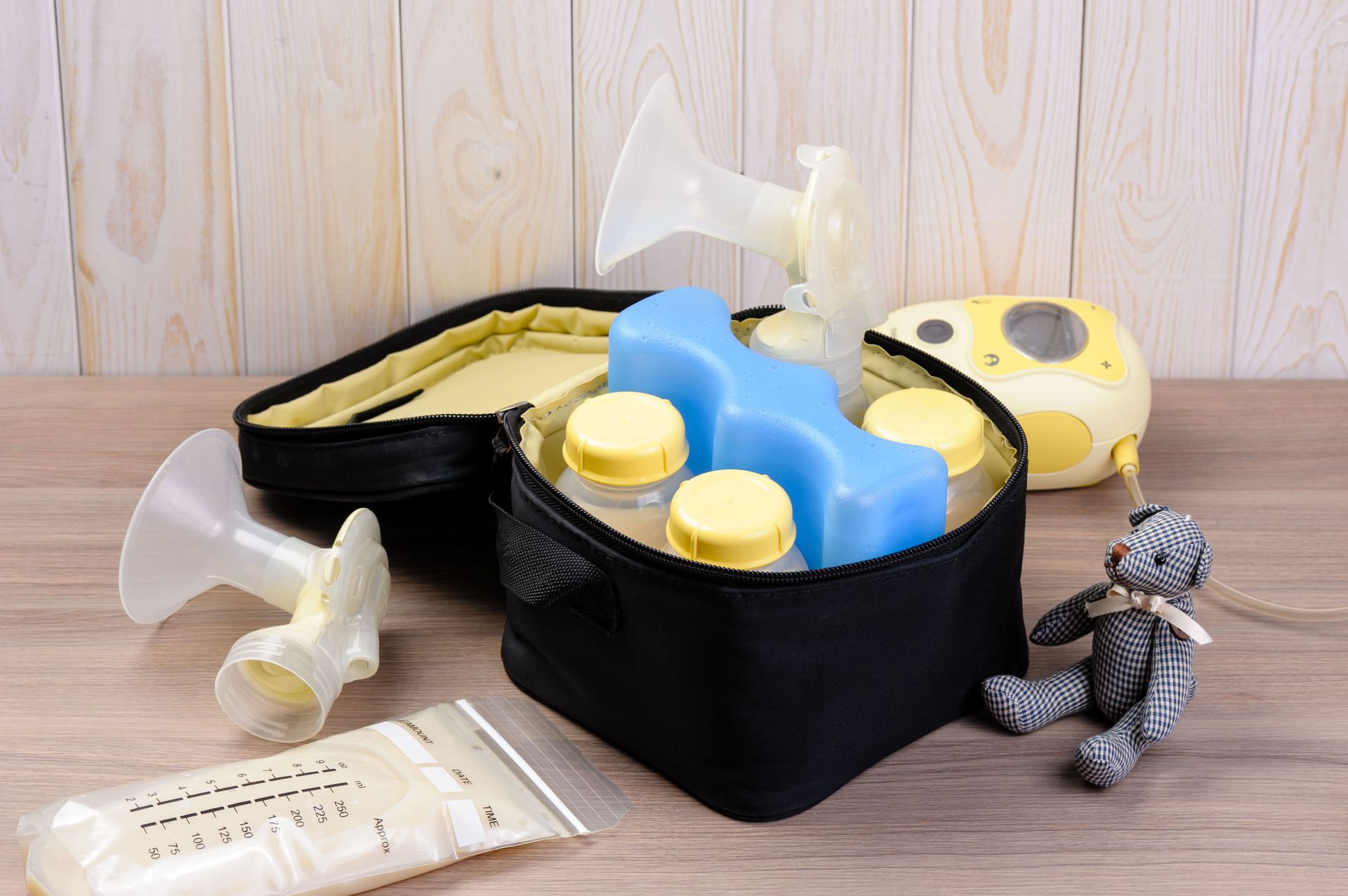 Breast pump and accessories including bottles, cooler pack, and a toy bear on wooden surface.
