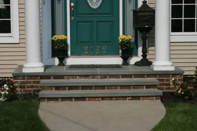 Green front door with white columns, brick steps, and potted yellow flowers at the entrance