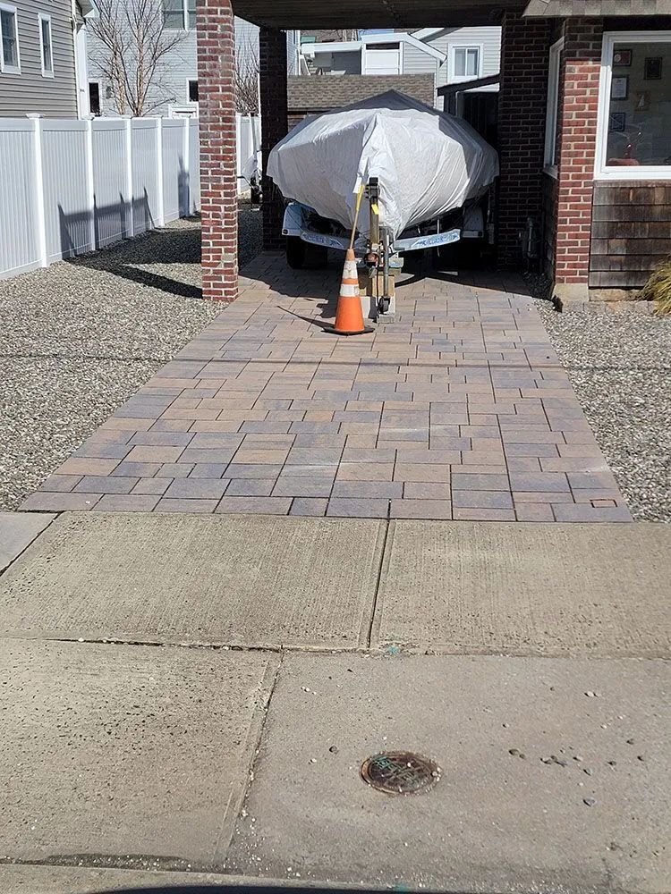 Covered car parked under a carport at the end of a brick-paved driveway with an orange cone in front.