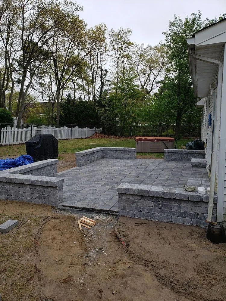 Backyard patio under construction with gray stone retaining walls and pavers, near a house and trees
