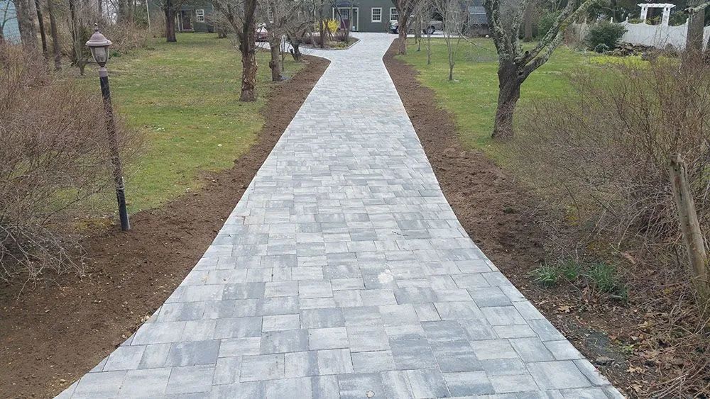 Stone walkway leading through a landscaped yard toward a house, with grass and bare shrubs on both sides