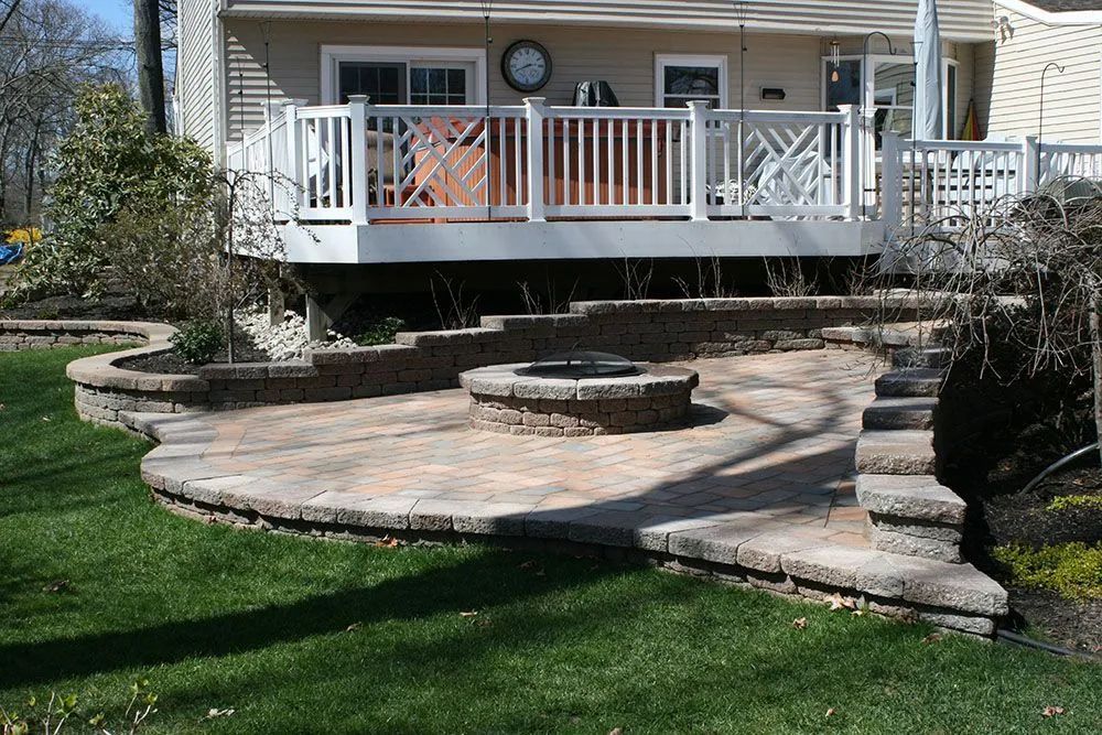 Backyard patio with stone steps and a raised wooden deck with white railing and chairs.