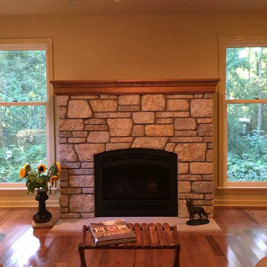 Stone fireplace between two tall windows in a sunlit living room, with flowers and a small table in front.