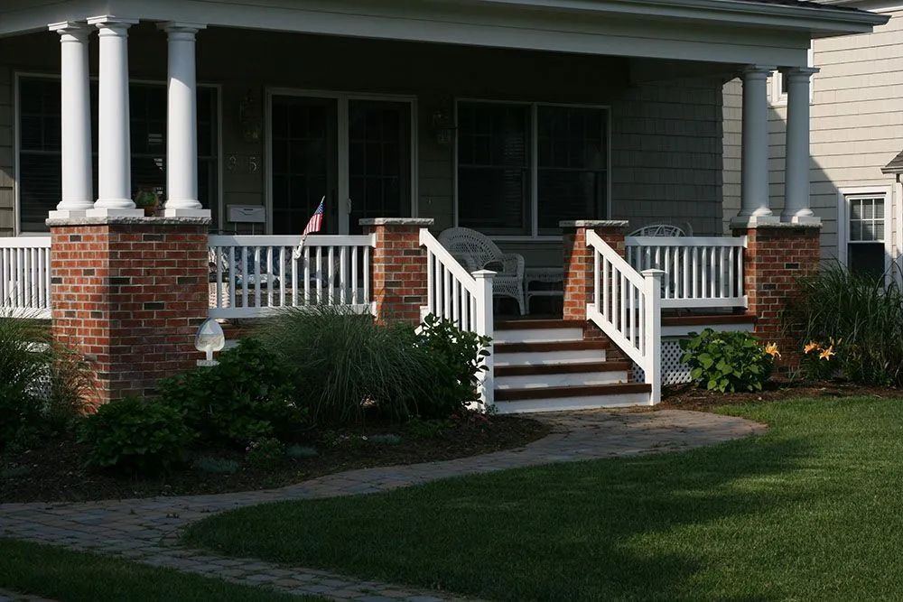 Front porch with white railings and brick columns on a house, with steps leading down to a lawn.