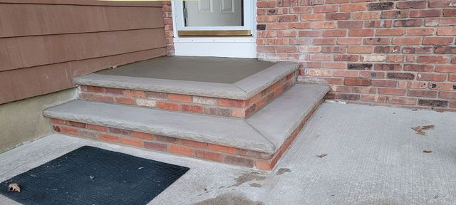 Concrete front steps with a small porch, brick walls, and a doormat by the entrance.