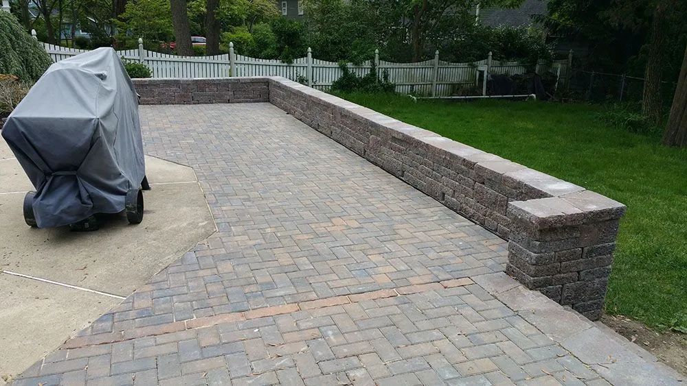 Paved patio with a covered grill, stone retaining wall, and green lawn in the background